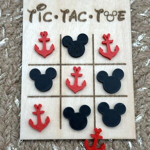 A wooden tic-tac-toe game board with red anchor and black Mouse head game pieces inspired for your next cruise. The board has the words 'Tic-Tac-Toe' written on it. These would be perfect Fish Extender or Pixie Dust gifts for your upcoming trip!