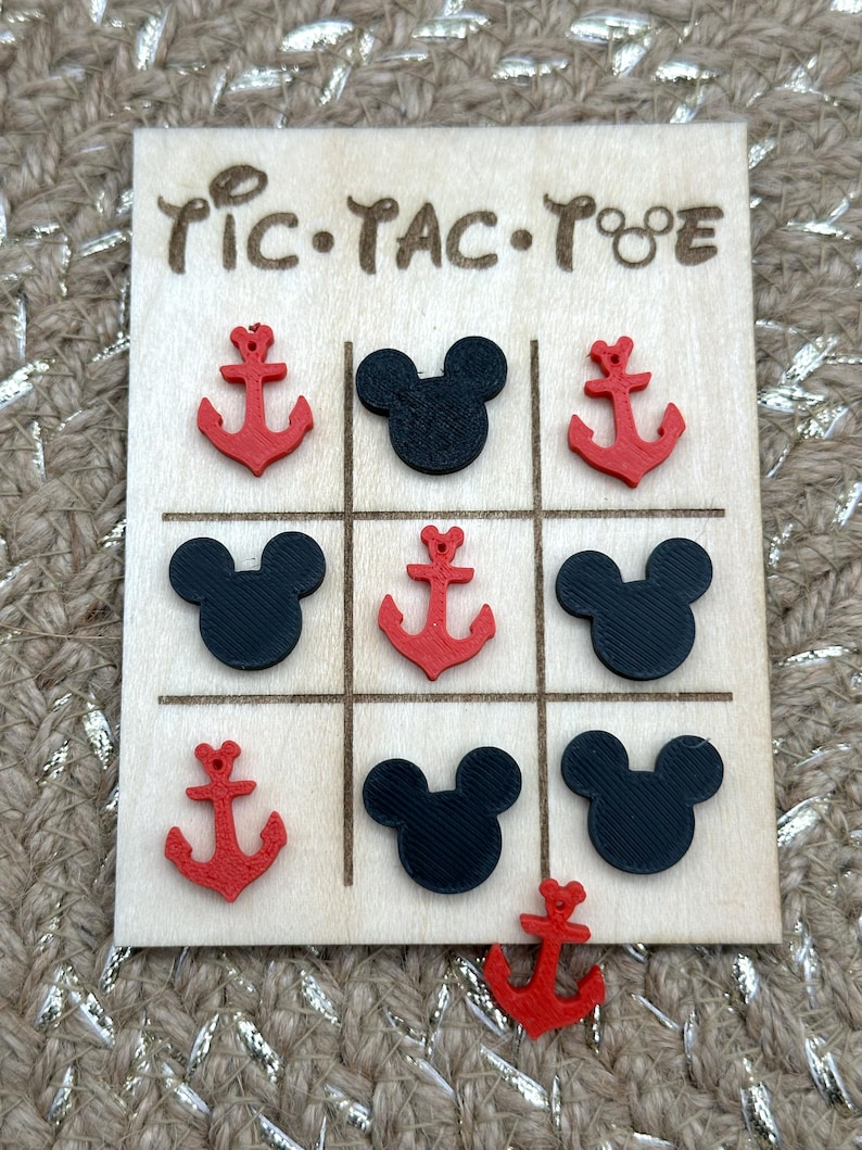 A wooden tic-tac-toe game board with red anchor and black Mouse head game pieces inspired for your next cruise. The board has the words 'Tic-Tac-Toe' written on it. These would be perfect Fish Extender or Pixie Dust gifts for your upcoming trip!