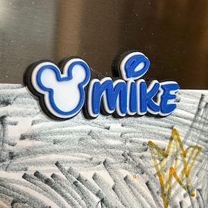 A blue and white 3D printed name tag with the name 'MIKE' and a Mouse head. The font of the name is in a mouse inspired font. Each magnet will have a Mouse head on the left of the name.