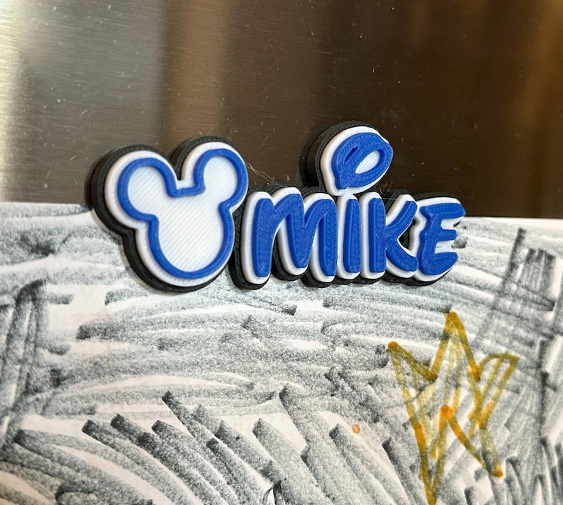 A blue and white 3D printed name tag with the name 'MIKE' and a Mouse head. The font of the name is in a mouse inspired font. Each magnet will have a Mouse head on the left of the name.