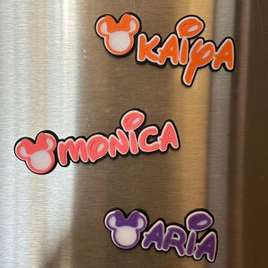 Three personalized name magnets with a Mouse head design in the girl version with a bow. The magnets are in orange, pink and purple. Each magnet will have 3 layers: black bottom layer, white middle layer and your choice of color for the top layer.