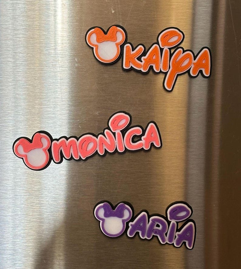 Three personalized name magnets with a Mouse head design in the girl version with a bow. The magnets are in orange, pink and purple. Each magnet will have 3 layers: black bottom layer, white middle layer and your choice of color for the top layer.