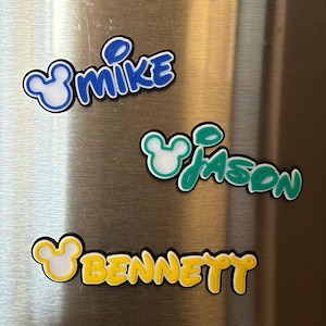 Three personalized name magnets with a Mouse head design in the boy version. The magnets are in blue, green, and yellow. Each magnet will have 3 layers: black bottom layer, white middle layer and your choice of color for the top layer.