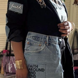 May include: A black blazer with denim accents, featuring patches with the word "faith" and a denim skirt with the text "FAITH AROUND AND FIND OUT." The outfit includes gold jewelry and a decorative brooch.