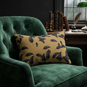 May include: A rectangular throw cushion with a mustard yellow background and a dark grey leaf pattern, resting on a deep green tufted velvet armchair. The cushion features a repeating botanical motif of scattered leaves across its surface.