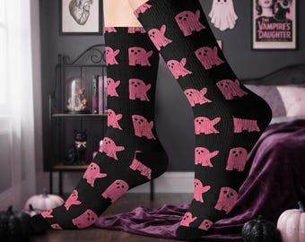 May include: A pair of black crew socks featuring a repeating pattern of small, pink, ghost-like figures with simple facial features. The socks have a ribbed texture and extend to the mid-calf, showcasing a consistent graphic print across the fabric.
