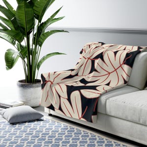 May include: A cream-coloured throw blanket featuring a repeating pattern of large, stylised leaves in cream and red against a dark navy background. The blanket is draped over a white sofa. A large potted plant is in the background.