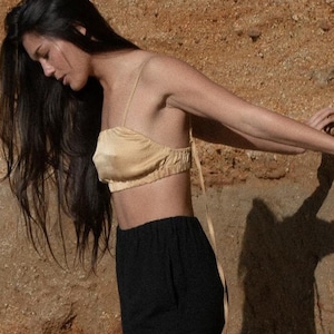 May include: A woman wearing a black top and black pants stands in front of a brown rock wall. Her hair is long and dark, and she is looking down.