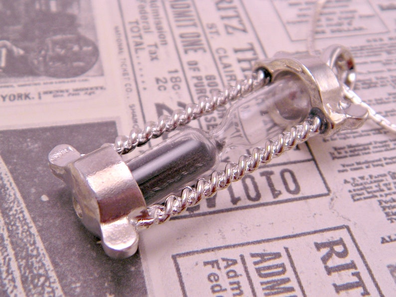 May include: A silver hourglass necklace with a black sand timer and a twisted silver chain. The necklace is on a white and black patterned surface.