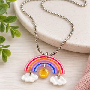 May include: A silver-toned ball chain necklace featuring a translucent, multi-colored rainbow pendant. The rainbow has red, orange, purple, and blue stripes, with a small yellow circular charm hanging in the center and two white clouds attached.