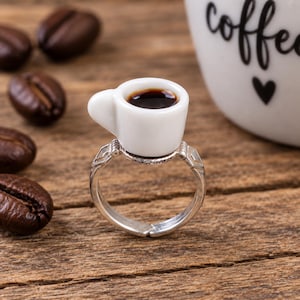 May include: A silver-toned metal ring featuring a miniature white ceramic coffee cup as the centerpiece. The tiny cup is filled with dark brown liquid, and the ring band has a textured, geometric design with an adjustable open back.