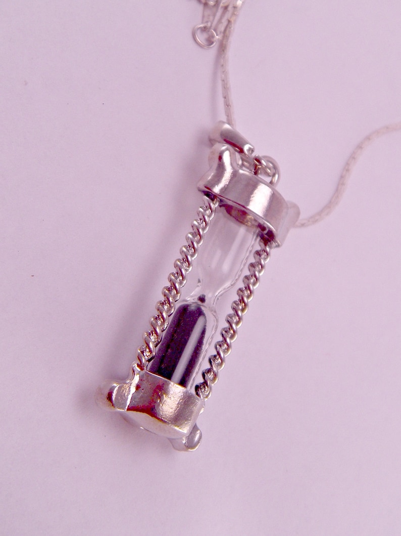May include: A silver necklace with an hourglass pendant. The hourglass is filled with black sand and has a clear glass tube.