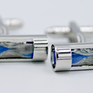 May include: A pair of silver cufflinks with a unique hourglass design. Each cufflink features a clear tube with blue and silver sand, creating a visual representation of time. The cufflinks have a polished finish.