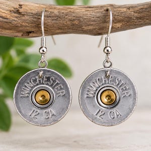 May include: A pair of silver-toned drop earrings featuring circular Winchester 12 GA shotgun shell casings. Each earring has a central brass-colored primer and is suspended from a simple silver-toned metal hook wire for pierced ears.