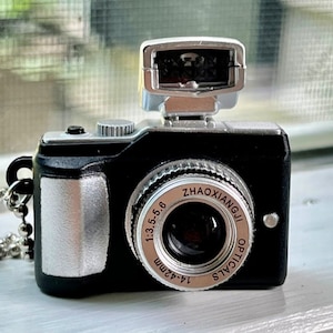 May include: A black and silver miniature camera keychain with a silver flash and a lens that reads "14-42mm 1:3.5-5.6 ZHAOXIANGJI OPTICALS".