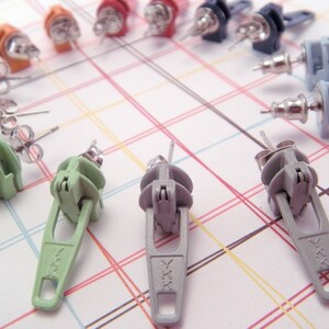May include: A set of 10 zipper pull earrings in various colors, including black, green, gray, and orange. The earrings are made of metal and have a zipper pull design.