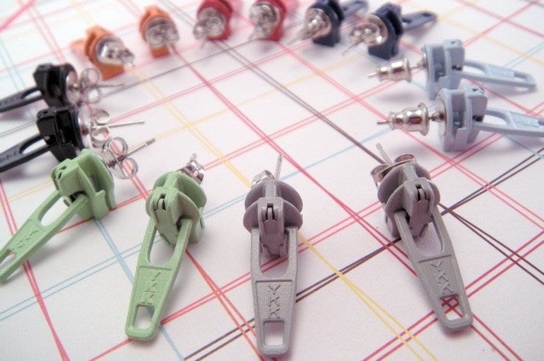 May include: A set of 10 zipper pull earrings in various colors, including black, green, gray, and orange. The earrings are made of metal and have a zipper pull design.
