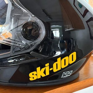 May include: Black motorcycle helmet with a yellow "ski-doo" logo on the side. The helmet has a clear visor and a black M-Forge logo.