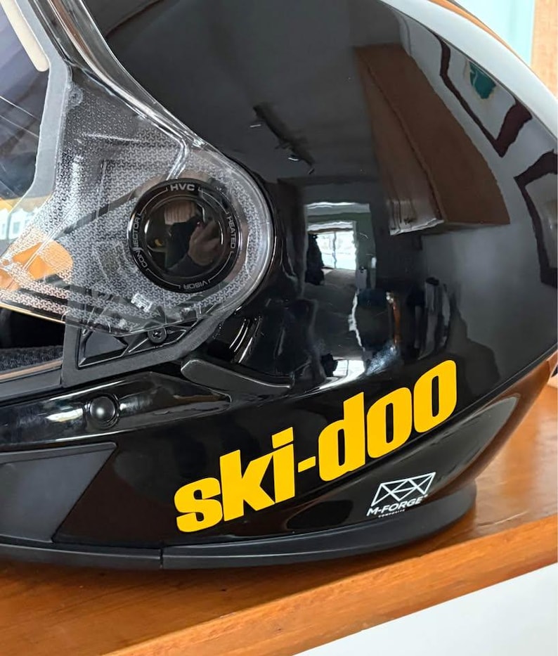 May include: Black motorcycle helmet with a yellow "ski-doo" logo on the side. The helmet has a clear visor and a black M-Forge logo.