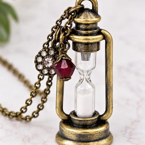 May include: A necklace featuring a miniature hourglass pendant encased in an antique brass-toned metal frame. The chain is adorned with a circular charm set with clear crystals and a faceted deep red bead, with white sand inside the glass hourglass.