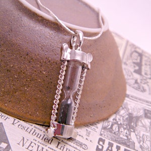 May include: A silver chain necklace with a silver hourglass pendant. The hourglass is filled with black sand and is suspended from the chain by a silver ring.