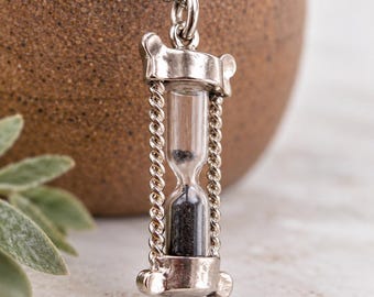 May include: A silver-toned pendant shaped like a miniature hourglass, featuring a clear glass center filled with fine black sand. The frame is constructed with twisted metal pillars and smooth end caps, suspended from a silver-toned ball chain necklace.