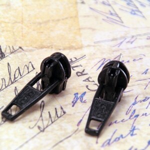 May include: Two black zipper pulls, commonly used for clothing or bags, are shown on a piece of paper with handwritten notes.