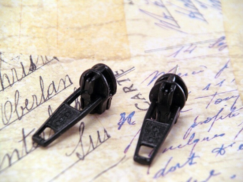 May include: Two black zipper pulls, commonly used for clothing or bags, are shown on a piece of paper with handwritten notes.