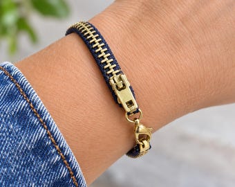 Puede incluir: Una pulsera fabricada con una cremallera de tela vaquera azul oscuro con dientes metálicos en tono dorado y un cursor a juego. La pieza cuenta con un cierre de mosquetón en tono dorado para una sujeción segura alrededor de la muñeca.
