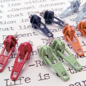 May include: A set of ten zipper earrings in various colors, including red, orange, green, blue, and gray. The earrings are displayed on a white surface with printed text.