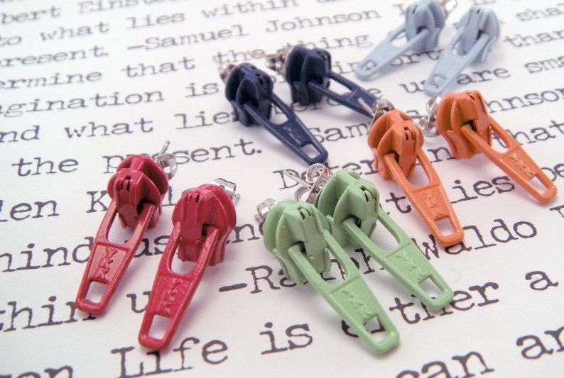 May include: A set of ten zipper earrings in various colors, including red, orange, green, blue, and gray. The earrings are displayed on a white surface with printed text.