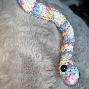 May include: A crocheted snake toy with a rainbow color pattern. The snake has black eyes and is made with a white yarn base.