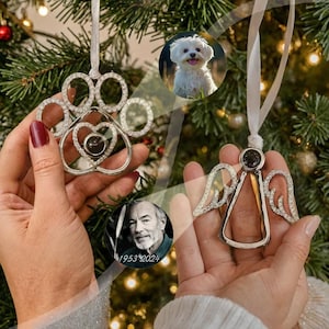 May include: Two silver Christmas ornaments with a photo insert. One ornament is a paw print with a heart in the center. The other ornament is an angel with wings. Both ornaments have rhinestones around the edges.