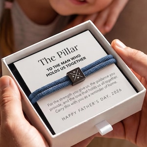 May include: A white gift box containing a blue bracelet with a dark square accent. The box has a card that reads "The Pillar TO THE MAN WHO HOLDS US TOGETHER" and "HAPPY FATHER'S DAY, 2026". A smiling child is in the background.