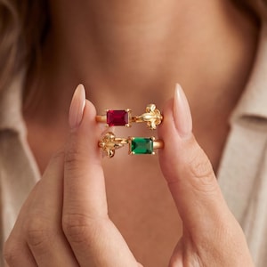 May include: Two gold rings with rectangular gemstones, one red and one green, are held up. The rings have a decorative floral design on the band. The background is a neutral color, highlighting the jewelry.