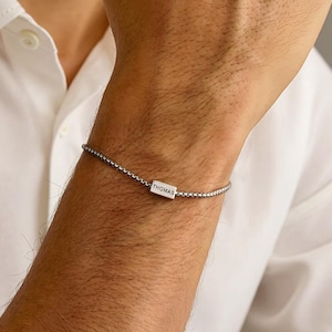 Peut inclure: Un bracelet argenté avec un petit charm rectangulaire. Le charm est gravé du nom "THOMAS". Le bracelet est porté au poignet, et la personne porte une chemise blanche boutonnée.