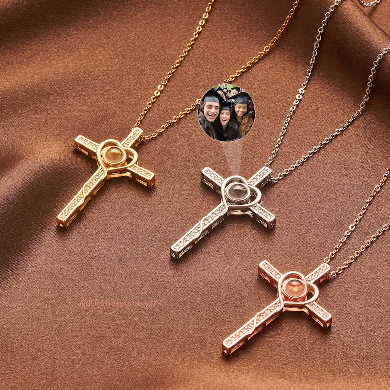 May include: Three gold, silver, and rose gold cross necklaces with heart-shaped pendants. Each pendant contains a small, round projection screen that displays a photo. The necklaces feature sparkling cubic zirconia stones.  The image displayed shows three smiling graduates. The watermark '@ElothejewelryUS' is visible.