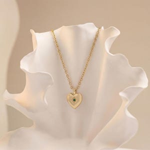 May include: A gold-colored heart-shaped pendant necklace with a small green gemstone in the center. The necklace has a delicate chain and is displayed against a white, petal-like background. The pendant is approximately 0.75 inches in width.