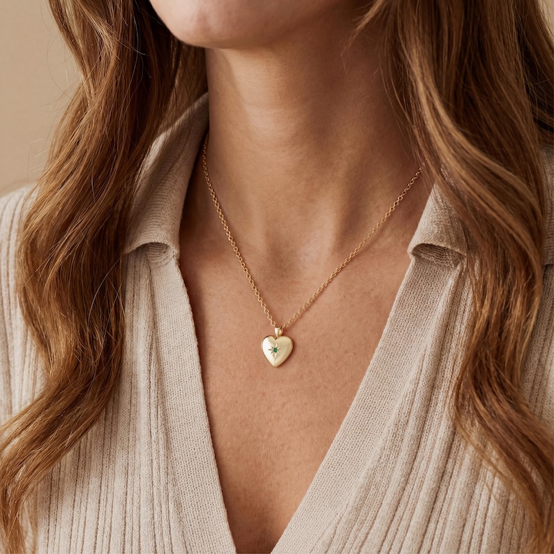 May include: A gold-colored heart-shaped pendant necklace with a small green gemstone in the center. The pendant hangs from a delicate gold chain. The necklace is worn against a beige top.
