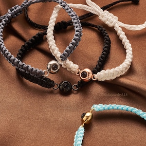 May include: Four braided bracelets with different colored cords and a silver, gold, and black eye charm. The bracelets are on a brown surface.  &copy;ElodiejewelryU5
