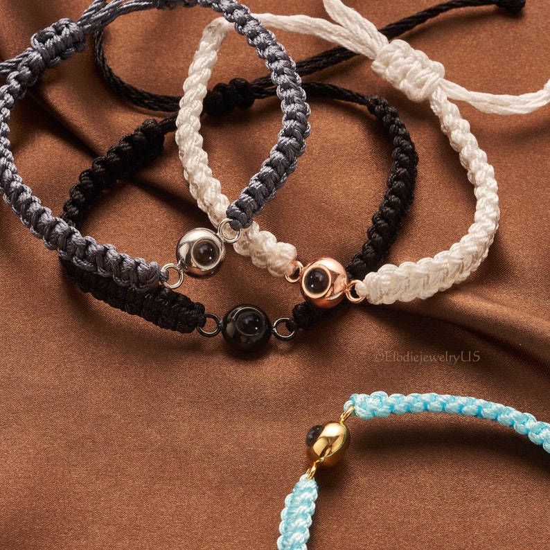 May include: Four braided bracelets with different colored cords and a silver, gold, and black eye charm. The bracelets are on a brown surface.  &copy;ElodiejewelryU5