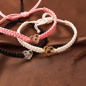 May include: Three friendship bracelets with heart charms. The bracelets are made of braided string in black, white, and pink. The heart charms have a clear crystal center.