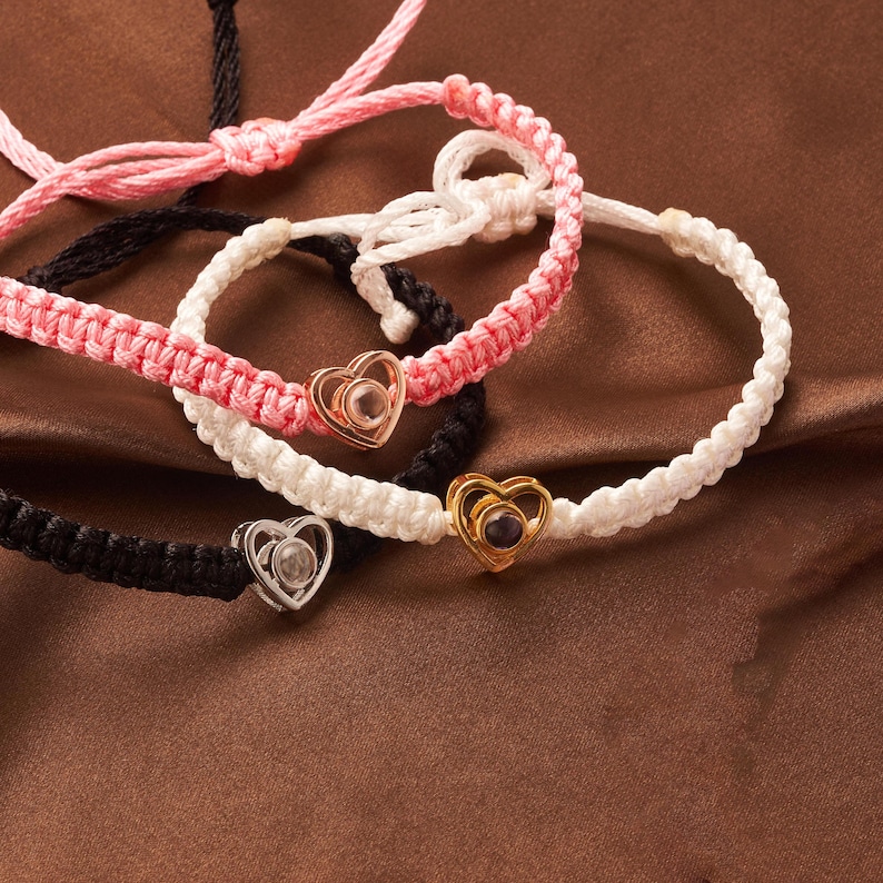 May include: Three friendship bracelets with heart charms. The bracelets are made of braided string in black, white, and pink. The heart charms have a clear crystal center.
