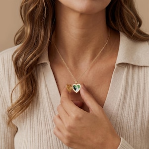 May include: A gold-colored heart-shaped locket necklace with a delicate chain. The locket is open, revealing a green and white image. The necklace is worn against a cream-colored top.