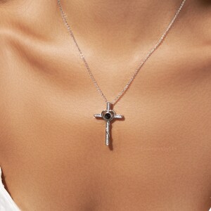 May include: Silver necklace with a cross pendant featuring a heart with a small black stone in the centre. The heart has a small inscription that is only visible when viewed from a specific angle.