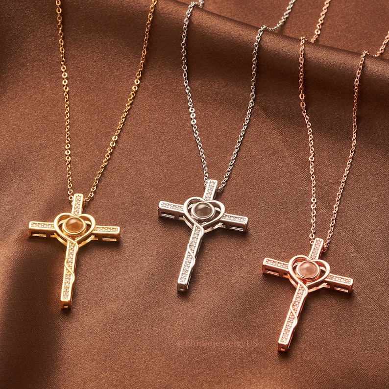 May include: Three silver cross necklaces with a heart-shaped pendant that projects an image when held up to a light source. The necklaces are available in gold, silver, and rose gold.