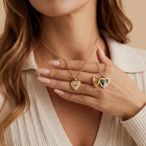 May include: Gold-colored heart-shaped locket necklaces. One locket is closed with a small green gemstone, and the other is open, revealing a dark interior. The necklaces are on delicate chains.