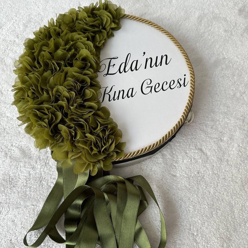 May include: A white tambourine with a gold trim and a green fabric flower design. The tambourine has the text "Eda'nun Kina Gecesi" written on it.