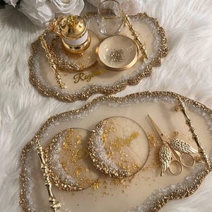 May include: Two gold-rimmed trays with white resin bases and gold accents. The larger tray has two round coasters with gold flakes and the name "Regina" written on them. The smaller tray has a gold-colored tea pot, a glass cup, and a small bowl with gold flakes. Both trays have a gold handle.