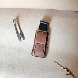 Może przedstawiać: Brązowe skórzane etui na magazynek z czarnym magazynkiem w środku. Etui ma klapę i jest przeszyte czarną nicią. Widoczne są również małe nożyczki i igła z czarną nicią.
